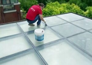 Worker applying transparent waterproof coating onto glass panel frame seams on a sunroom glass roof