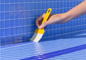 Applying clear liquid waterproofing sealant with a brush onto blue ceramic tile grout lines in a swimming pool basin